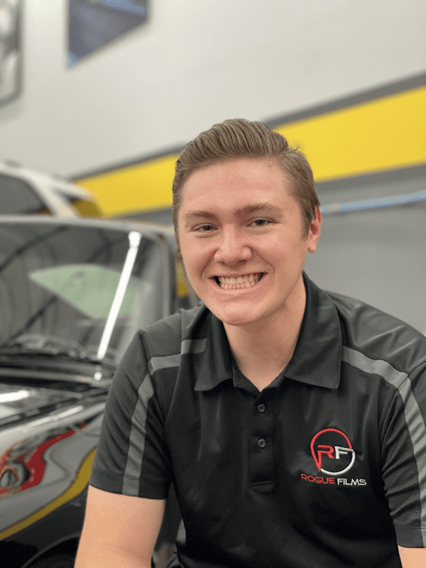 Young person with short hair smiling and wearing a black "Rogue Films" polo, standing in front of a yellow and black vehicle in what appears to be an indoor setting.