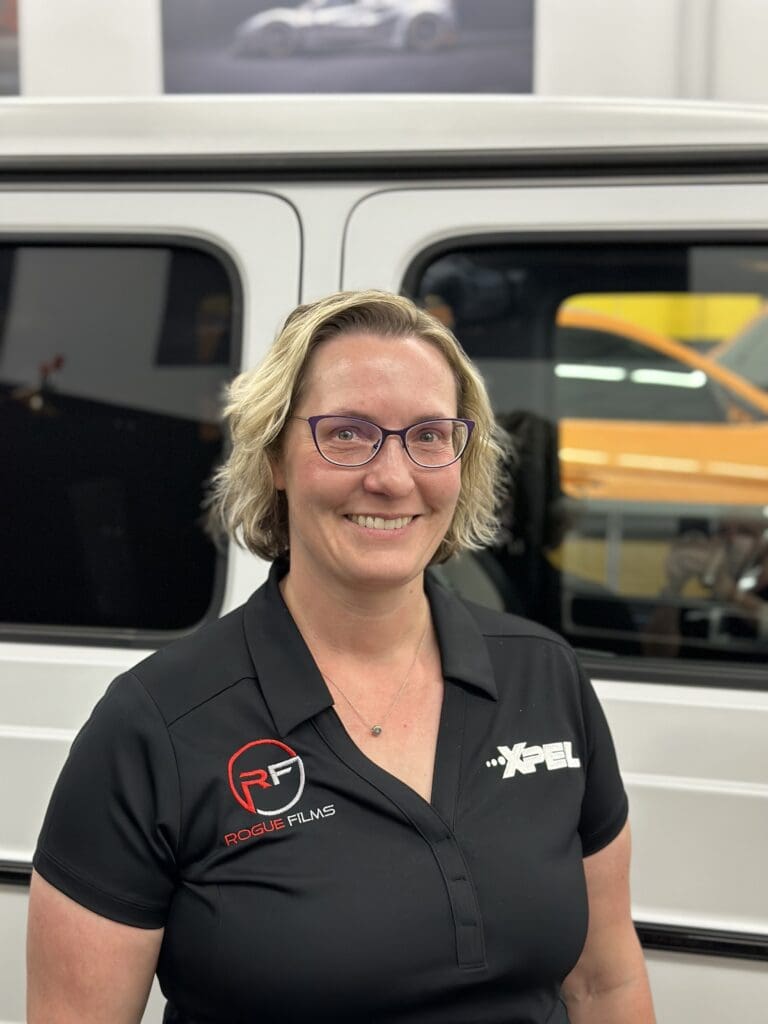 Woman with short blonde hair, glasses, and a black polo shirt with "Rogue Films" and "XPEL" logos, standing in front of a vehicle.