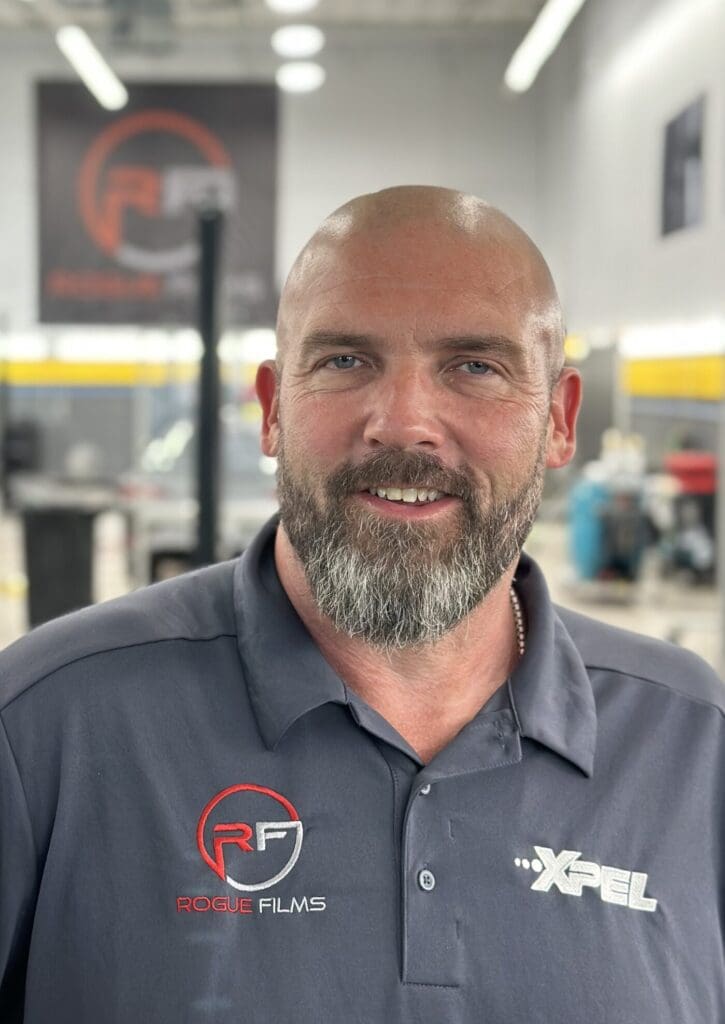 A man with a beard smiles while wearing a grey polo shirt with "Rogue Films" and "XPEL" logos. The background appears to be a workshop or industrial setting.