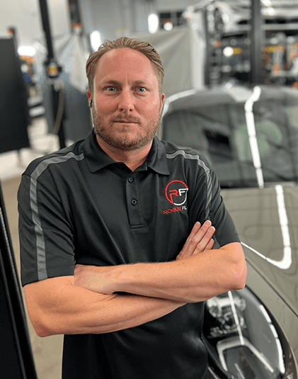 A person with short hair and a beard stands with arms crossed in a car workshop. They wear a black polo shirt with "RF" and "Rockin P L" logos. A vehicle is visible in the background.