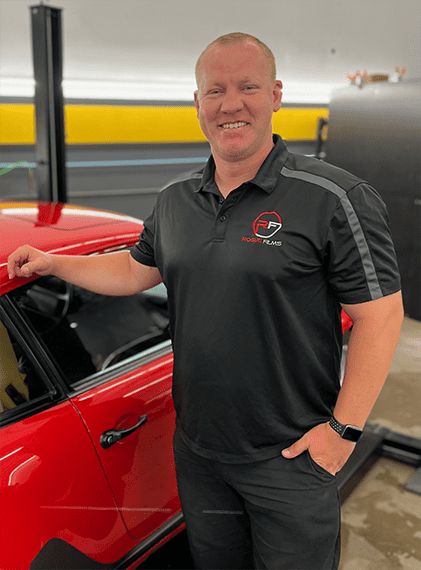 A man in a black polo shirt with a company logo stands next to a red car in a garage. The background includes a yellow and gray wall and automotive equipment.