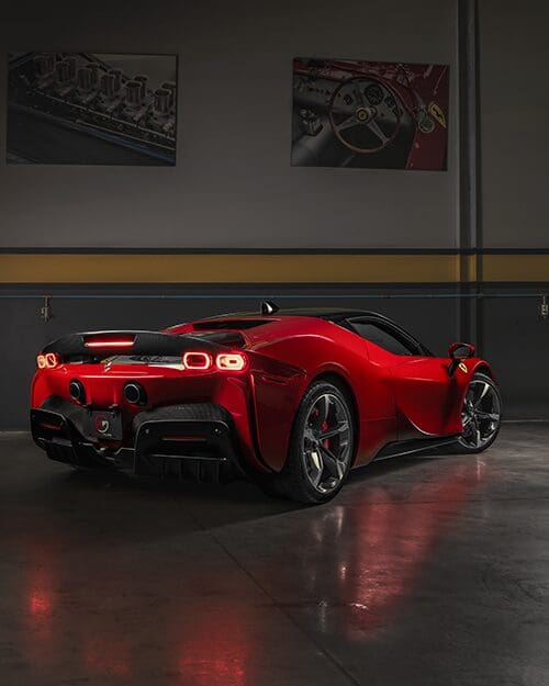 A sleek, red sports car is parked indoors, showcasing its rear design and aerodynamic features. Two automotive posters are on the wall in the background.