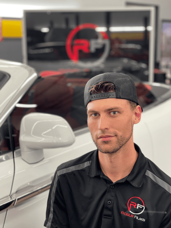 A man with a beard and a backward cap stands next to a white car, wearing a black Rogue Films polo shirt. A logo and the words "Rogue Films" are visible on a window in the background.