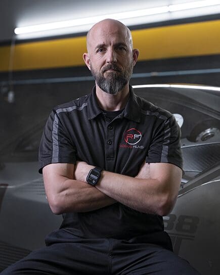 A man with a bald head and beard, wearing a black polo shirt, sits with his arms crossed in front of a car in a garage.