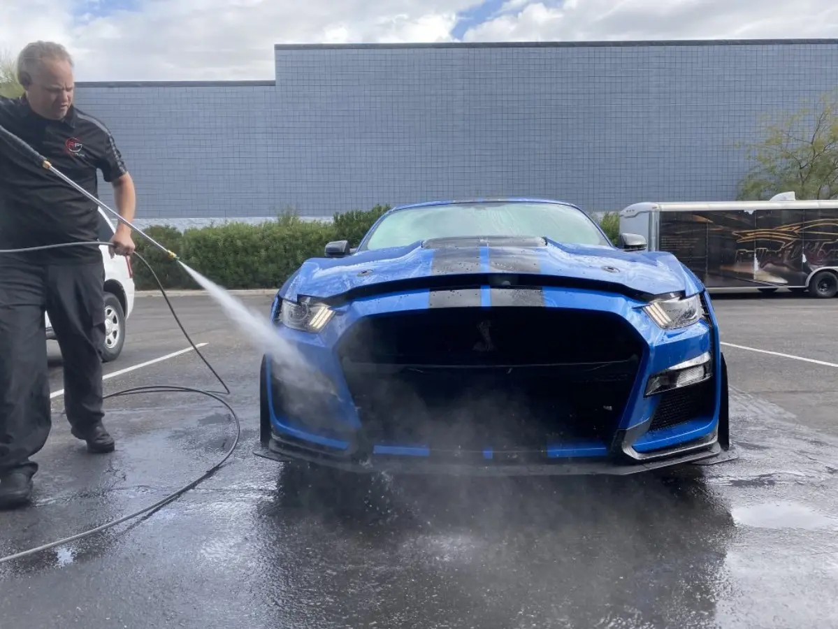 A person is using a pressure washer to clean the front of a blue sports car in a parking lot.