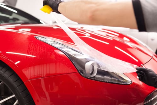 A person is applying a clear protective film to the headlight and front section of a red sports car.