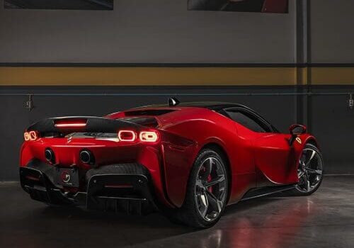 A sleek, red sports car is parked indoors, showcasing its rear design and aerodynamic features. Two automotive posters are on the wall in the background.