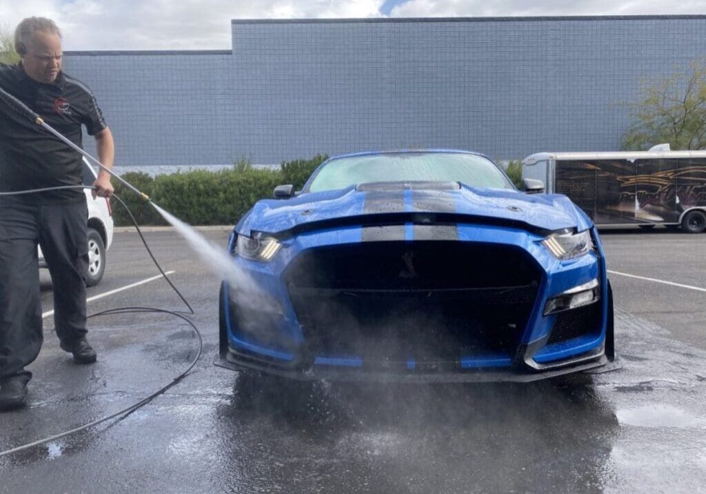 A person is using a pressure washer to clean the front of a blue sports car in a parking lot.