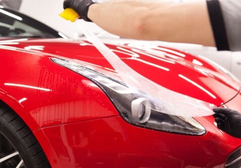 A person is applying a clear protective film to the headlight and front section of a red sports car.